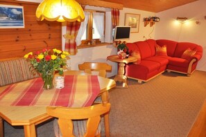 A cozy room with a wooden interior, a red sofa, a dining table with chairs, a lamp, and a TV on the wall. - Holiday apartment with 1 bedroom (Wallgau)