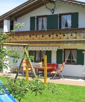 A two-story house with a balcony, a dining area with a table and chairs, and a garden with a swing set. - Holiday apartment with 1 bedroom (Wallgau)