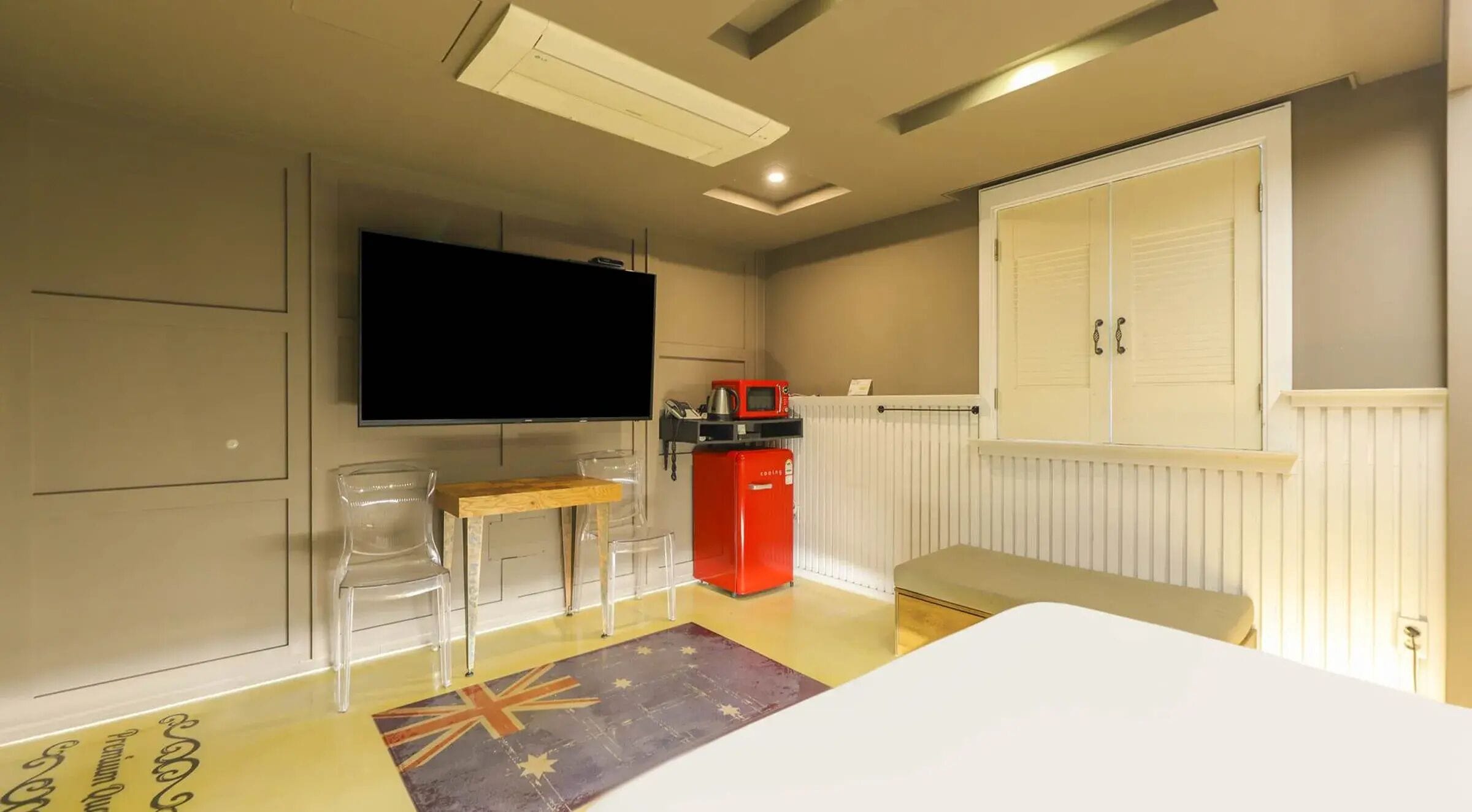 A room with a bed, a TV, a red appliance, and a window with a white blind.