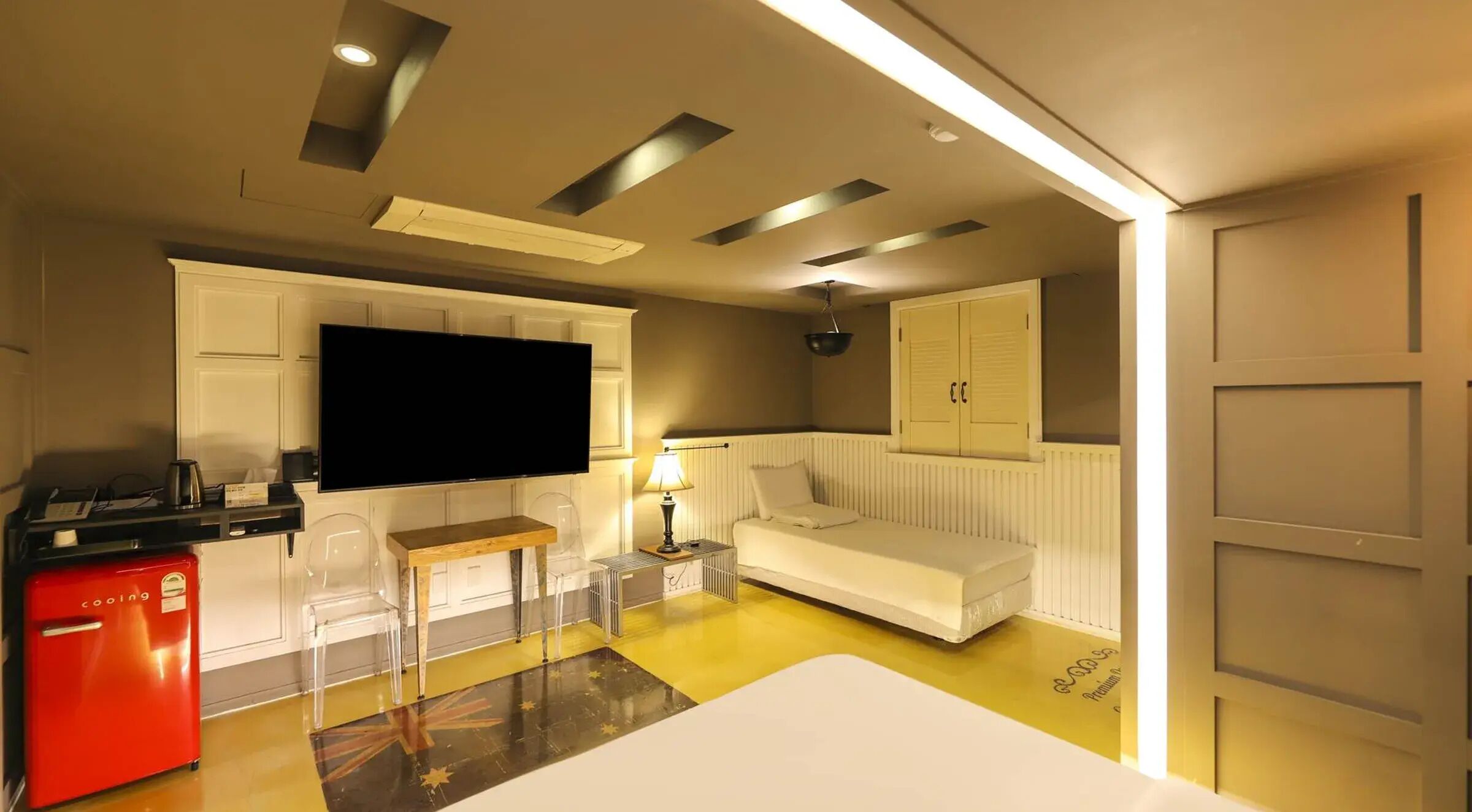 A compact hotel room with a flat-screen TV, a small kitchenette, a white sofa, a red fridge, and a yellow floor.