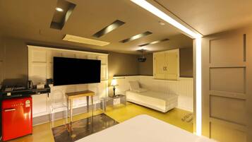 A compact hotel room with a flat-screen TV, a small kitchenette, a white sofa, a red fridge, and a yellow floor.