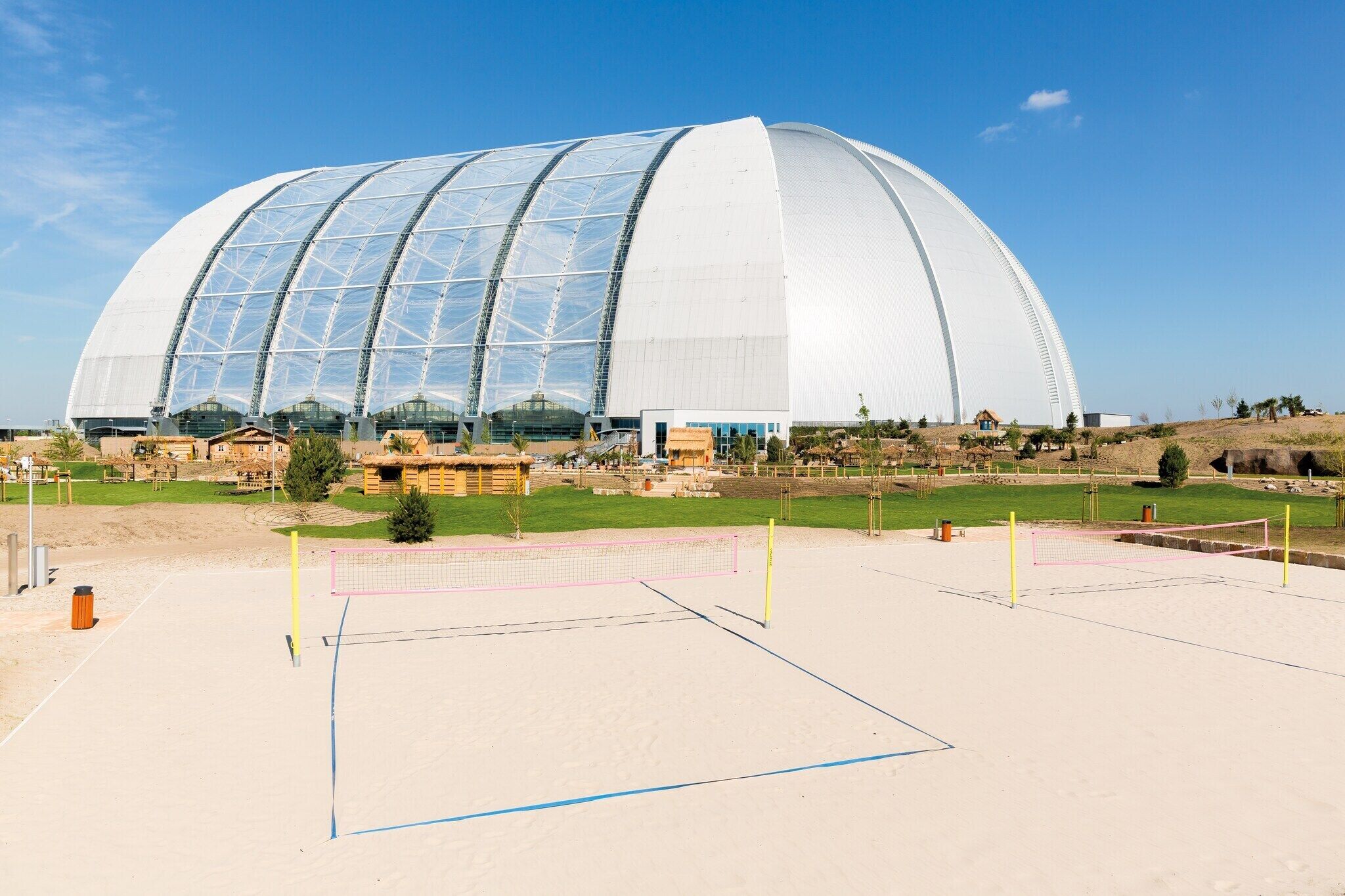 Un edifici gran de forma cònica i color blanc, amb un sostre transparent, envoltat d'una zona sorrenca i una xarxa de voleibol.