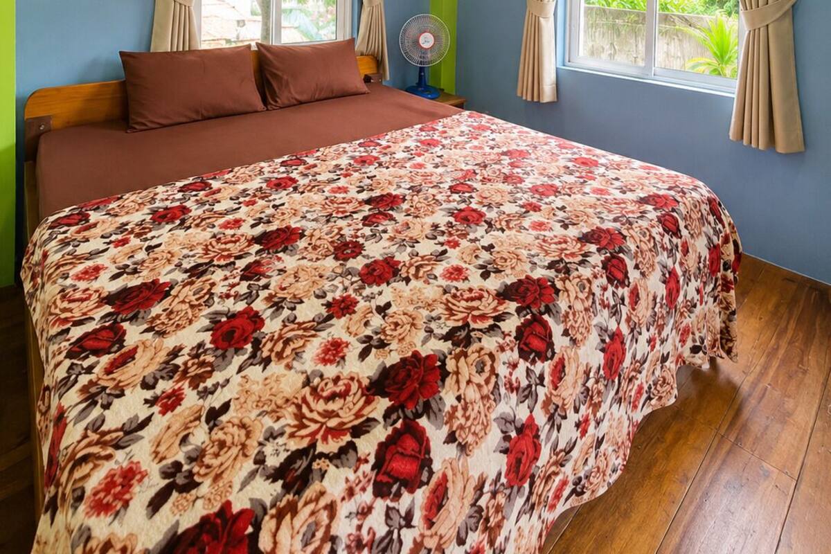 Ein Schlafzimmer mit floralem Bettbezug, zwei Fenstern mit Vorhängen, einem Deckenventilator und Holzboden.