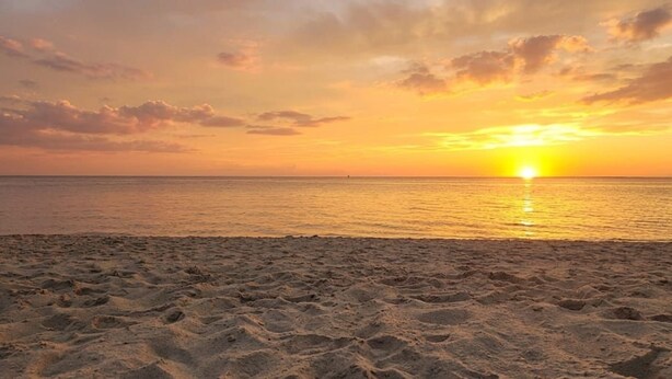 Una playa con huellas que van hacia el agua, una puesta de sol y un mar tranquilo.