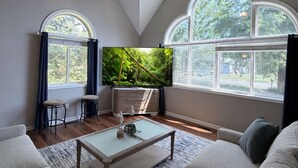 Una sala de estar con ventilador de techo, televisor de pantalla plana, mesa de centro y un amplio ventanal con cortinas.