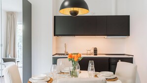 A dining area with a round table, chairs, a vase with flowers, and a modern kitchen in the background.