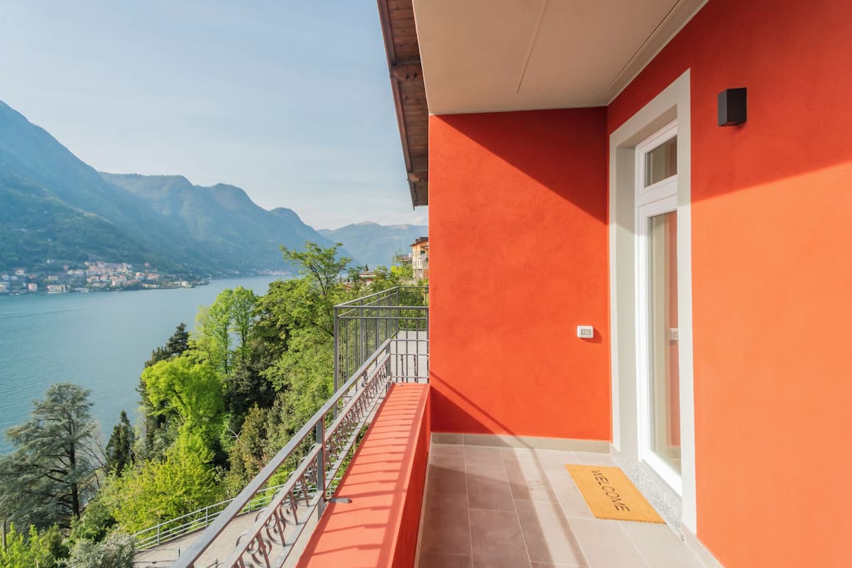 A balcony with a red wall, a metal railing, and a view of a lake and mountains.