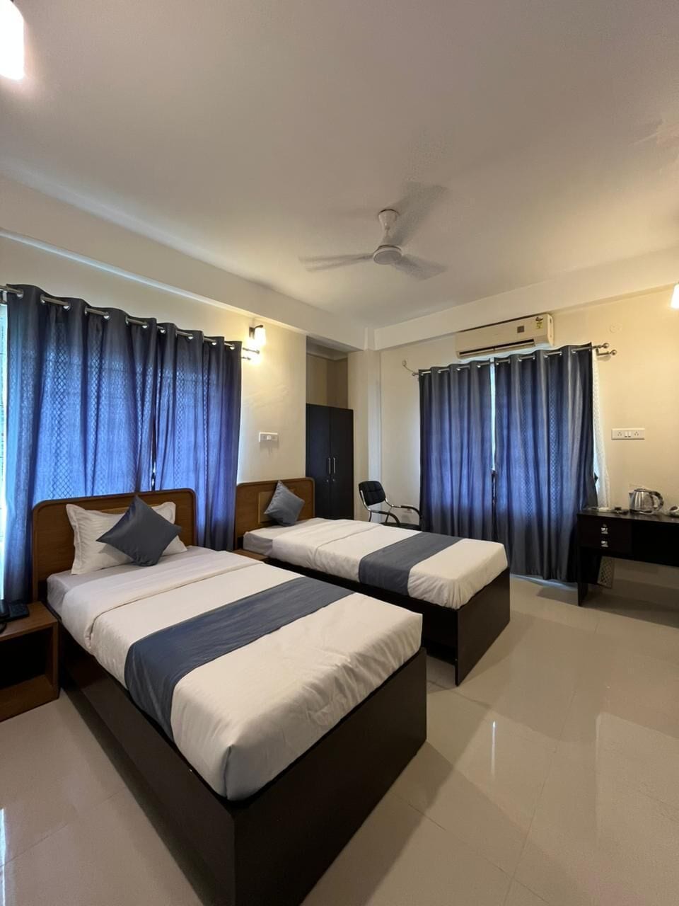 A hotel room with two beds, a ceiling fan, an air conditioning unit, a desk, and a chair.