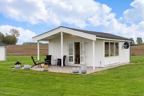 Ein eingeschossiges weißes Gebäude mit überdachter Terrasse, Sitzgelegenheiten im Freien und großen Fenstern.