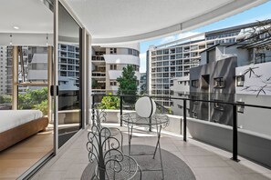 Eine moderne Wohnung mit Balkon, einem Essbereich mit Metallstühlen, einem Bett und Blick auf umliegende Gebäude.
