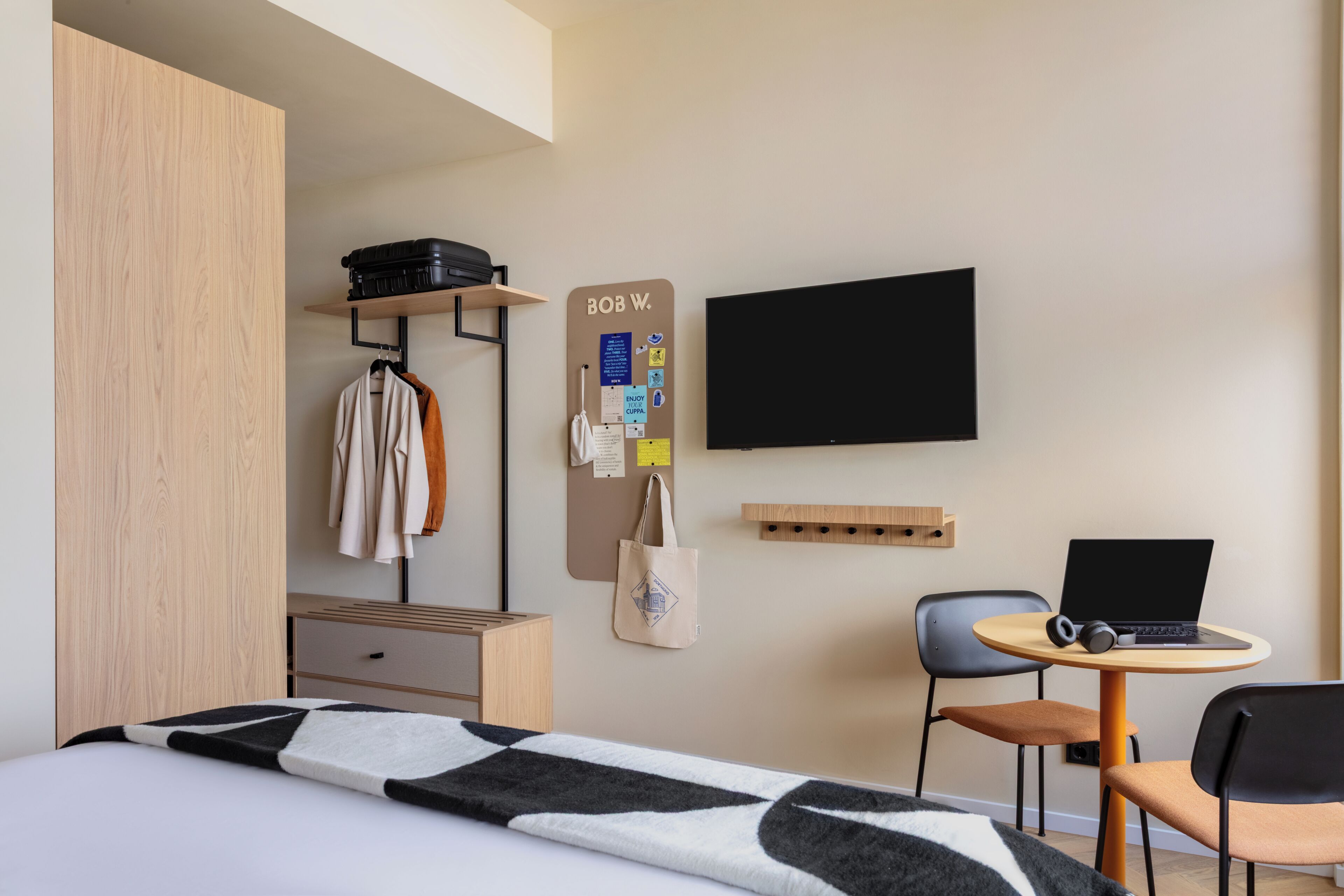A modern hotel room with a bed, a desk with a laptop, a chair, a TV, and a wardrobe.