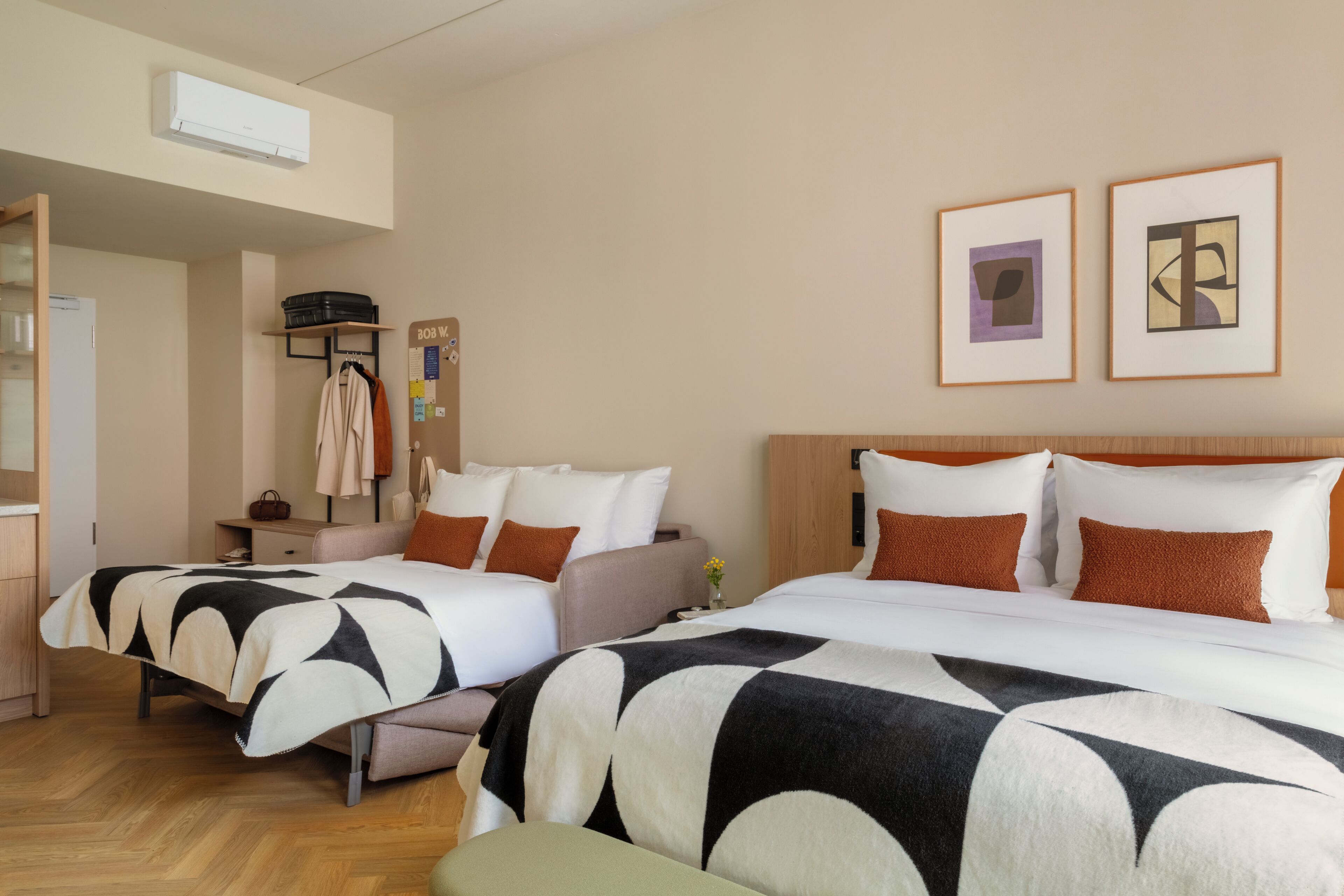 Une chambre d’hôtel avec deux lits, une tête de lit en bois, une table de chevet, une lampe, un climatiseur mural et des œuvres d’art encadrées.