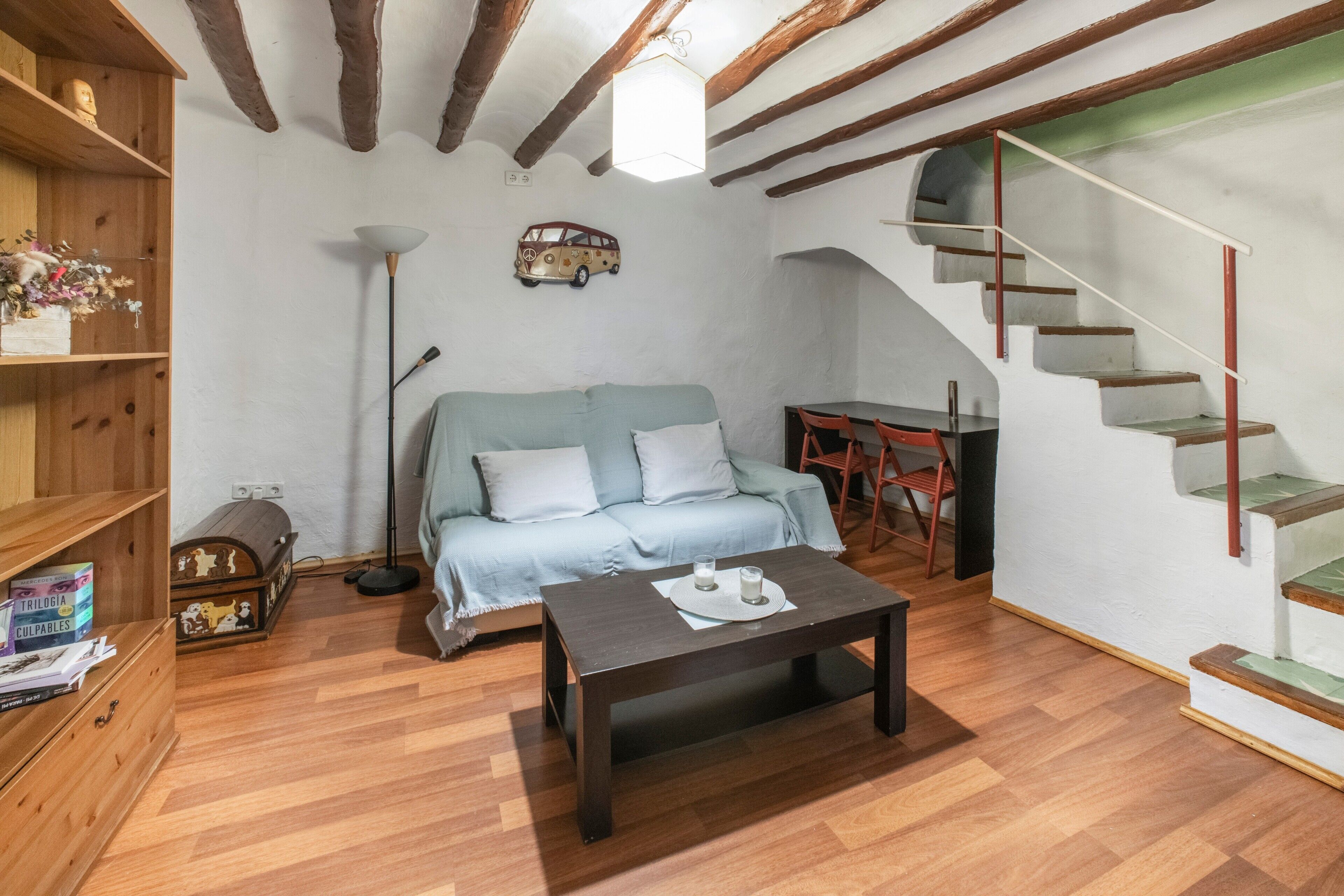 Una habitación con piso de madera, un sofá, una mesa de centro, una estantería, un escritorio y una escalera.