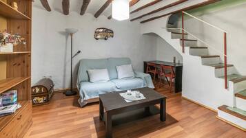 Una habitación con piso de madera, un sofá, una mesa de centro, una estantería, un escritorio y una escalera.