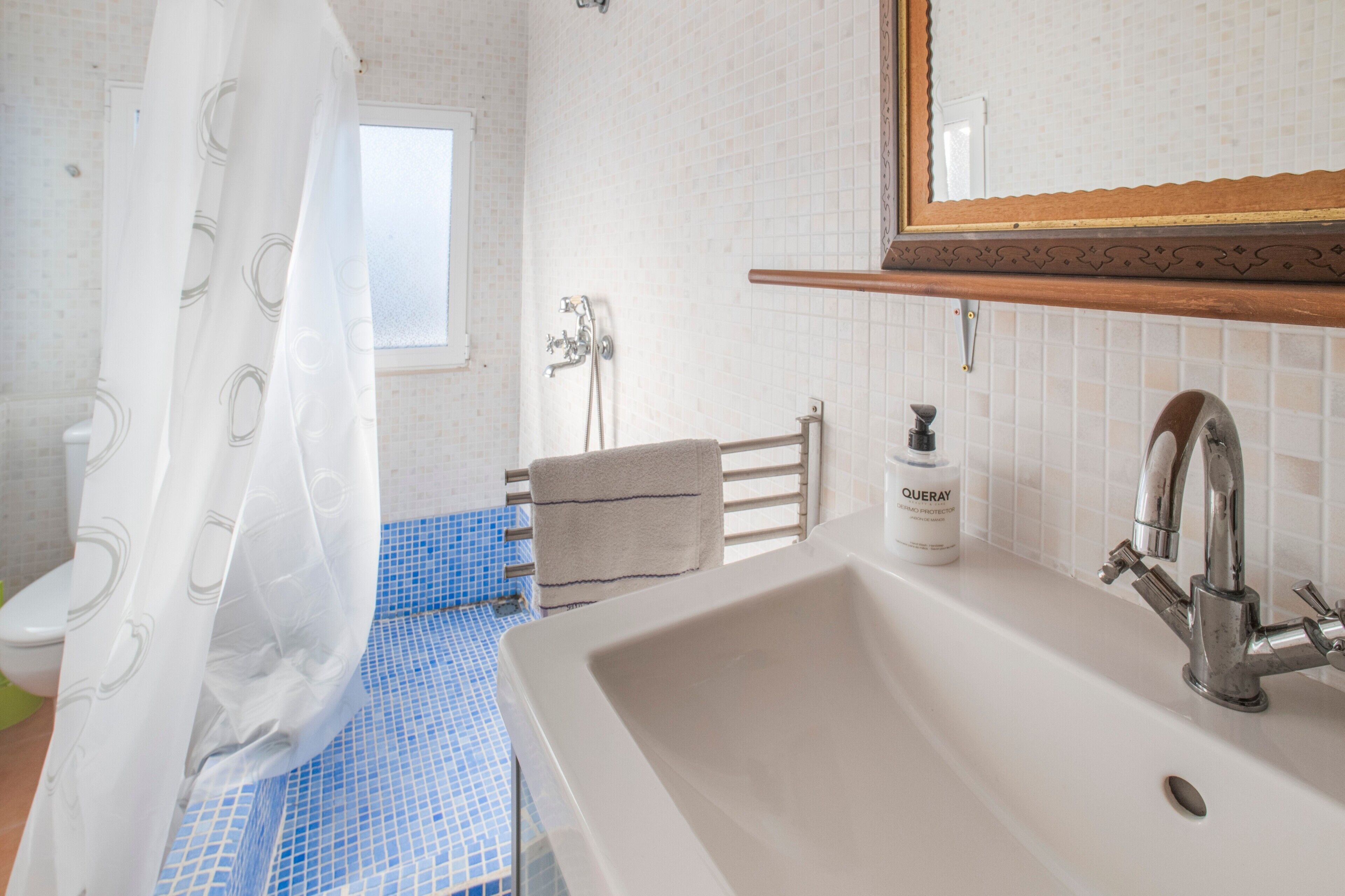 Un baño con lavamanos blanco, espejo, cortina de ducha, toallero y ventana.