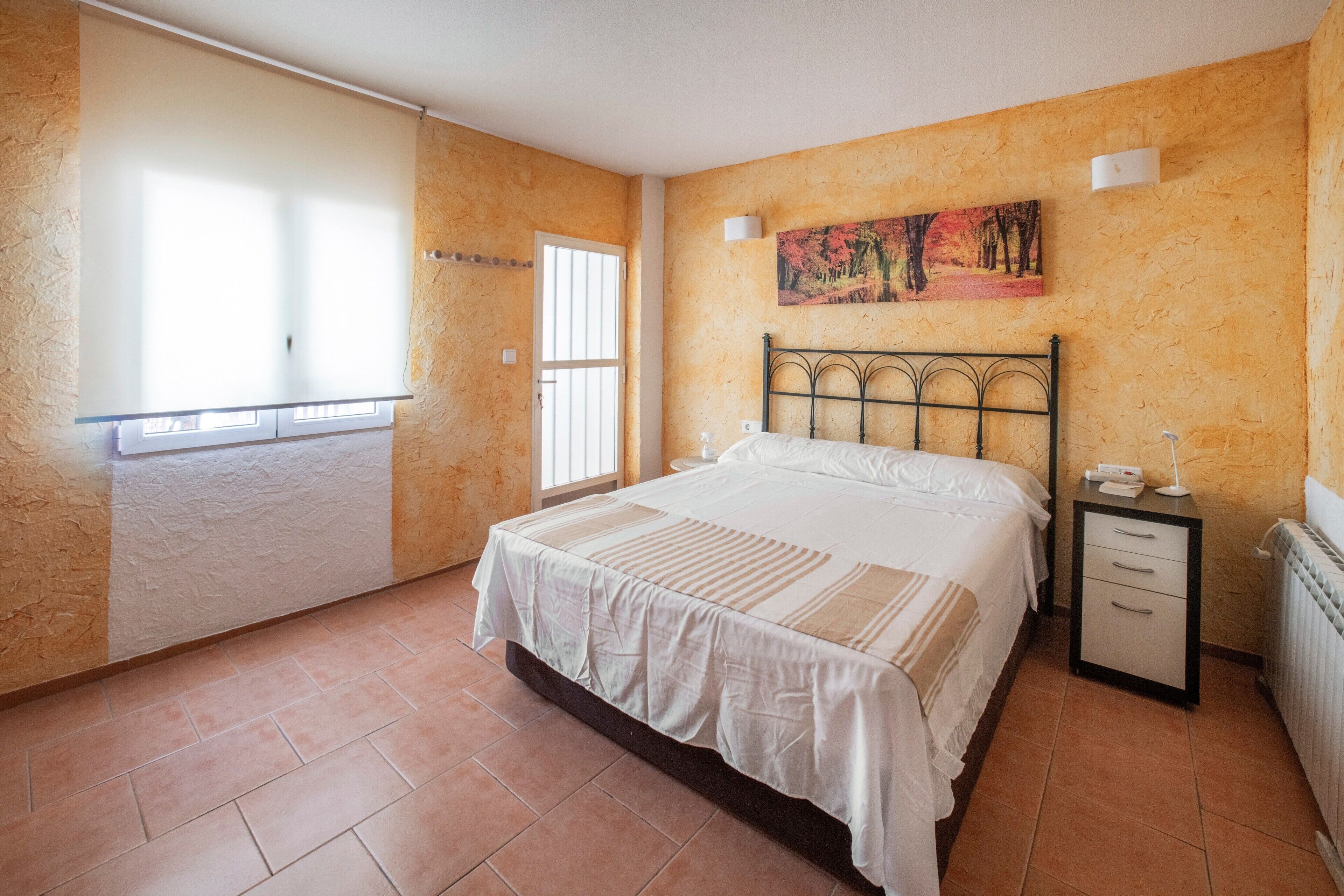 Un dormitorio con cama, mesita de noche, radiador, ventana con persianas y un cuadro en la pared.