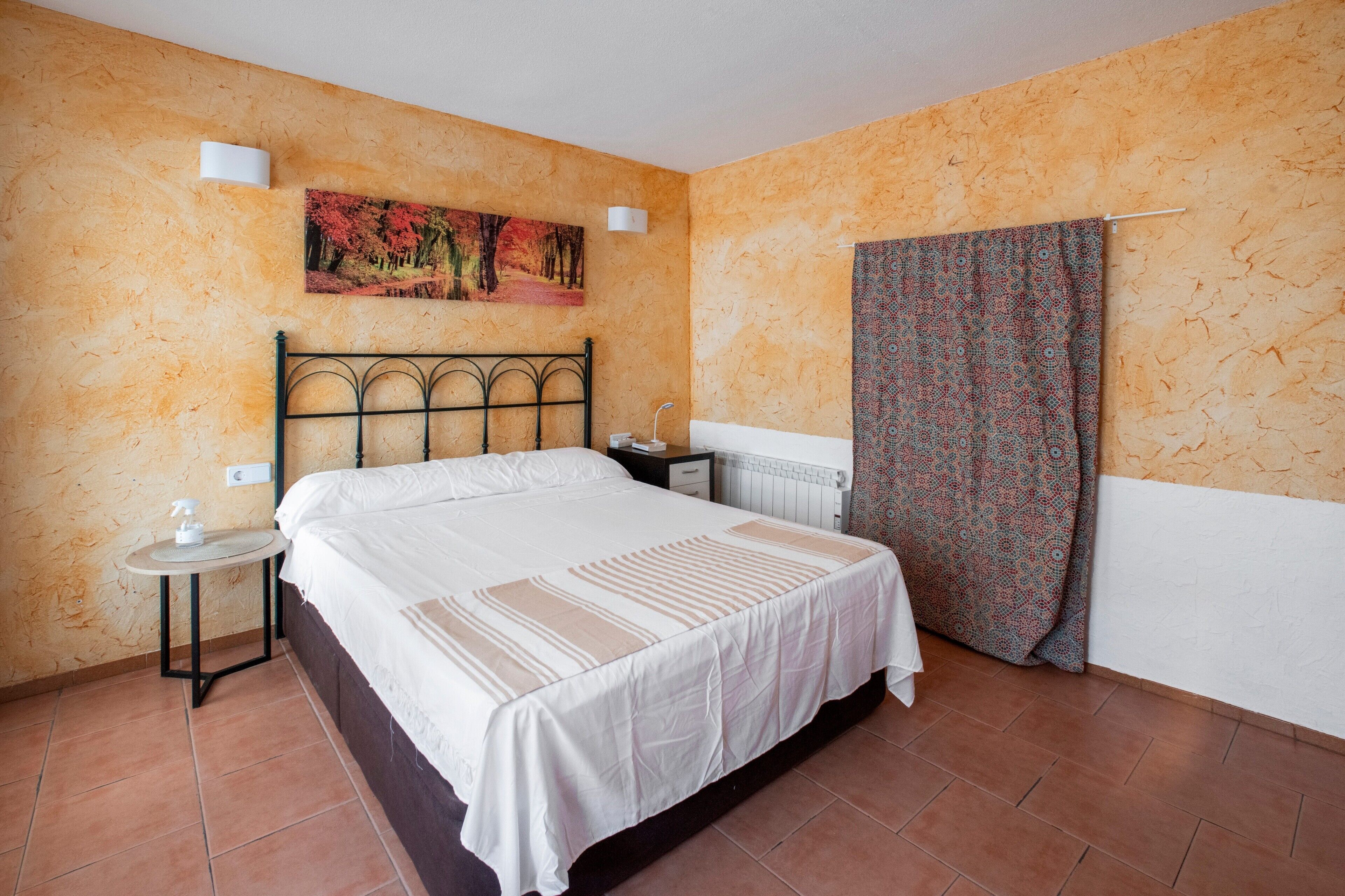 Un dormitorio con cama, mesita de noche, lámpara de pared, radiador y cortina estampada.