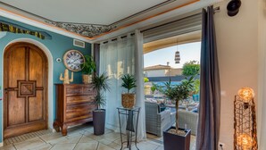 Ein Zimmer mit Holztür, einer Uhr, Topfpflanzen, einem Korbsessel und Blick auf einen Pool.