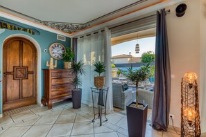 Quarto com porta de madeira, um relógio, plantas em vasos, um sofá de vime e vista para uma piscina.