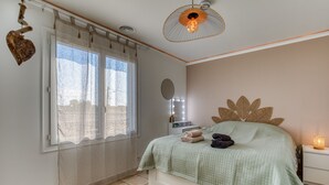 Ein Schlafzimmer mit Bett, einem Fenster mit Vorhängen, einem Nachttisch mit Lampe und einem an der Wand befestigten Spiegel.