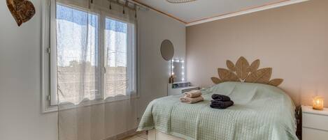 Une chambre à coucher avec un lit, une fenêtre avec des rideaux, une table de chevet avec une lampe et un miroir fixé au mur.