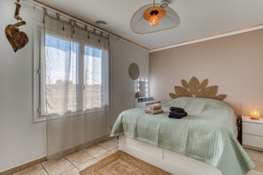 Quarto com cama, janela com cortinas, mesa de cabeceira com abajur e espelho fixado na parede.