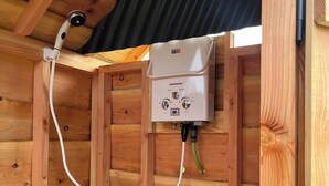 Calentador de agua instalado en una pared de madera, con un panel de control blanco, una manguera blanca y un techo corrugado negro arriba.