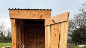 Una estructura de madera al aire libre con puerta, un banco pequeño y un baño.