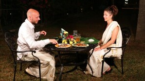 Two people seated at an outdoor table with plates of food, a glass of drink, and a small vase with flowers.