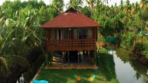 A wooden house with a red roof, surrounded by lush greenery and a water body.