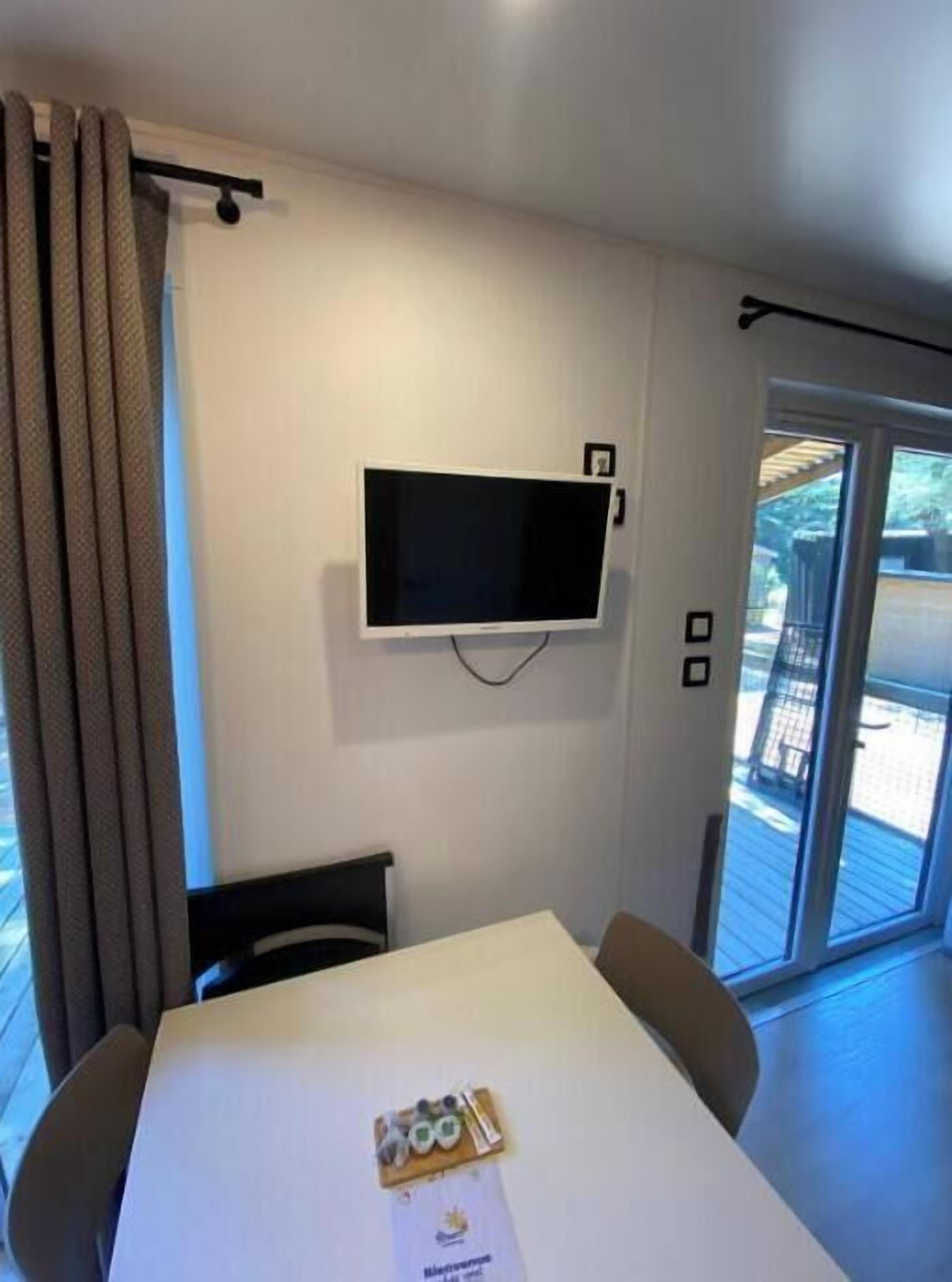 Una habitación con una televisión de pantalla plana montada en la pared, una mesa con sillas y vistas al exterior a través de una puerta de cristal.