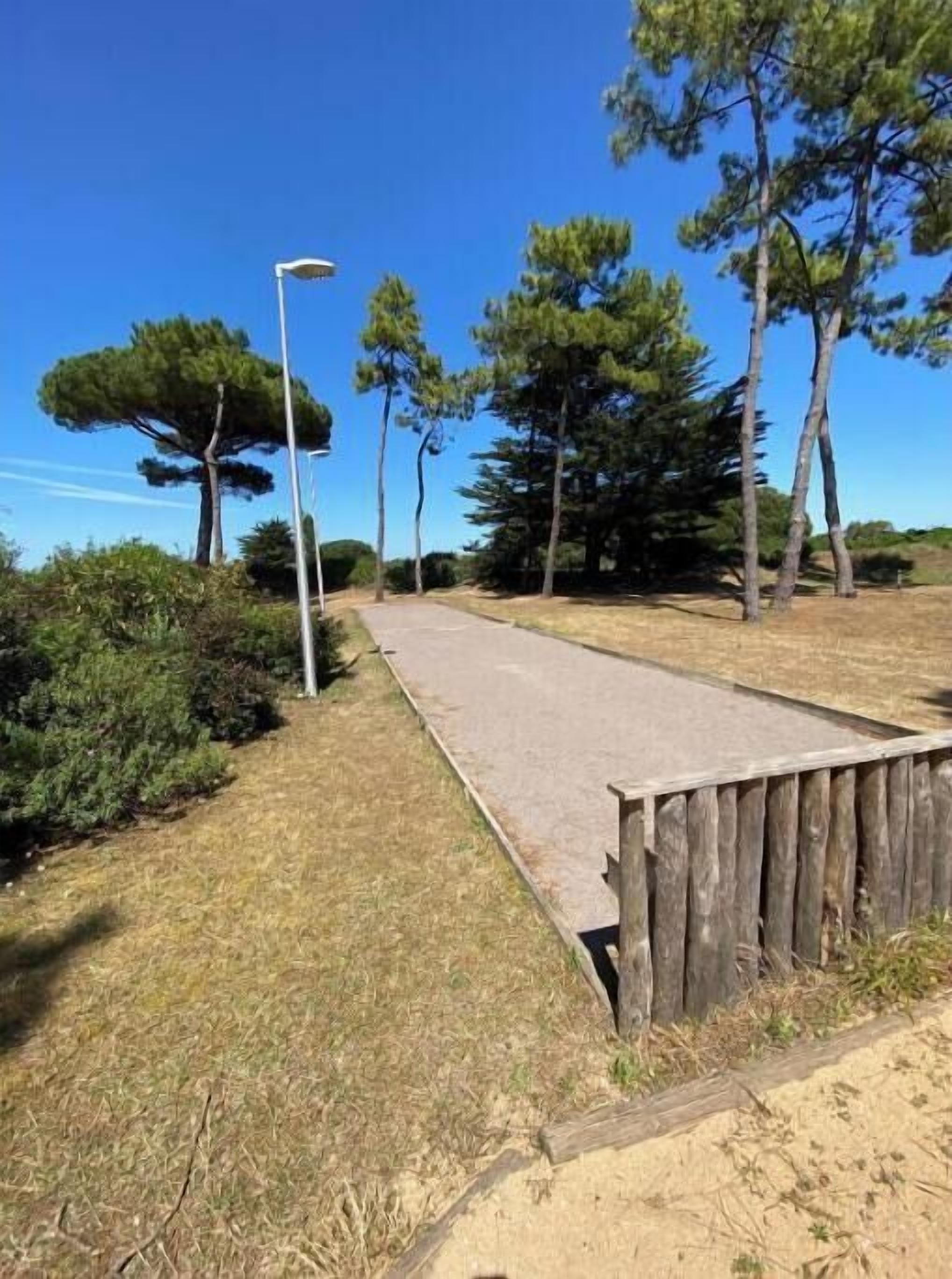 Un chemin pavé bordé d’une clôture en bois d’un côté, entouré de pins et surplombé d’un ciel bleu clair.