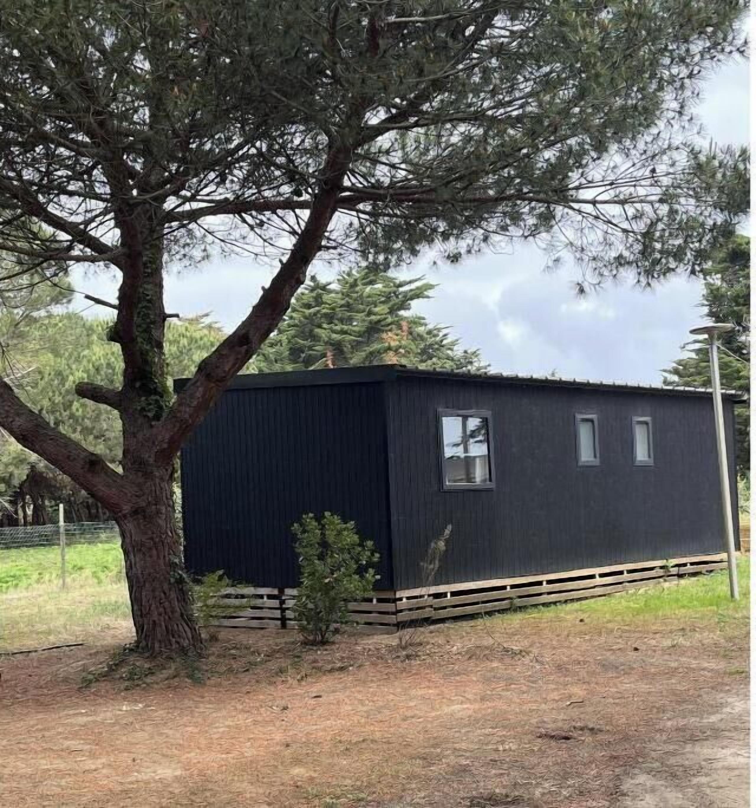 A small, dark-colored cabin with a single window and a door, surrounded by trees and a fenced area.