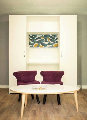 Una habitación blanca con piso de madera, una mesa blanca con dos sillas moradas y un armario blanco con repisas decoradas con motivos de hojas.