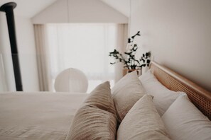 A bed with a wooden headboard, white bedding, and a view of a room with a large window and a plant.