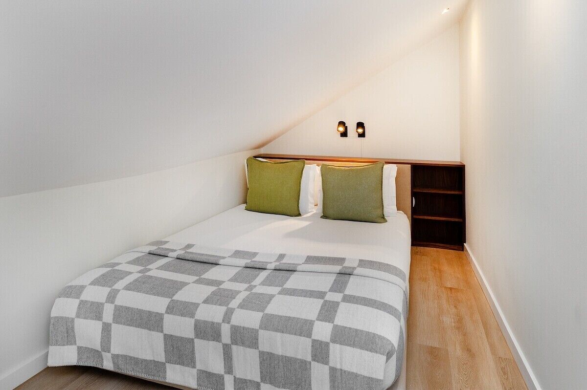 A bedroom with a bed, a checkered blanket, two pillows, a nightstand, and a wall-mounted light fixture.