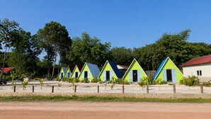 Una hilera de cabañas coloridas con techos triangulares, un camino de tierra en primer plano y árboles al fondo.
