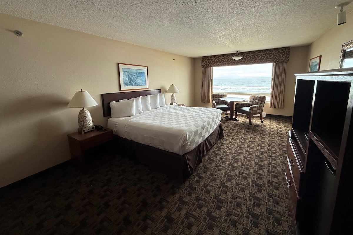 A hotel room with a bed, a desk with a lamp, a television, a chair, and a view of the ocean.