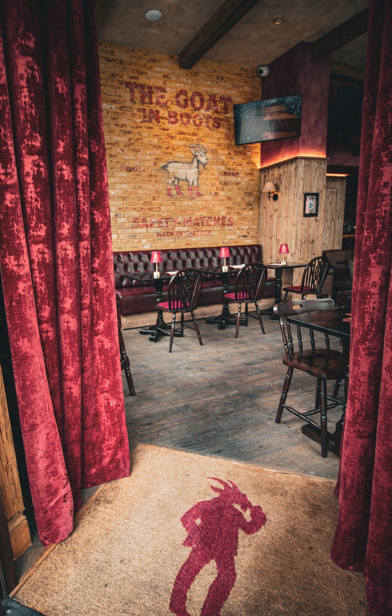 Interior acogedor de restaurante con pared de ladrillo, cortinas de terciopelo rojo, muebles de madera y un mural de una cabra.