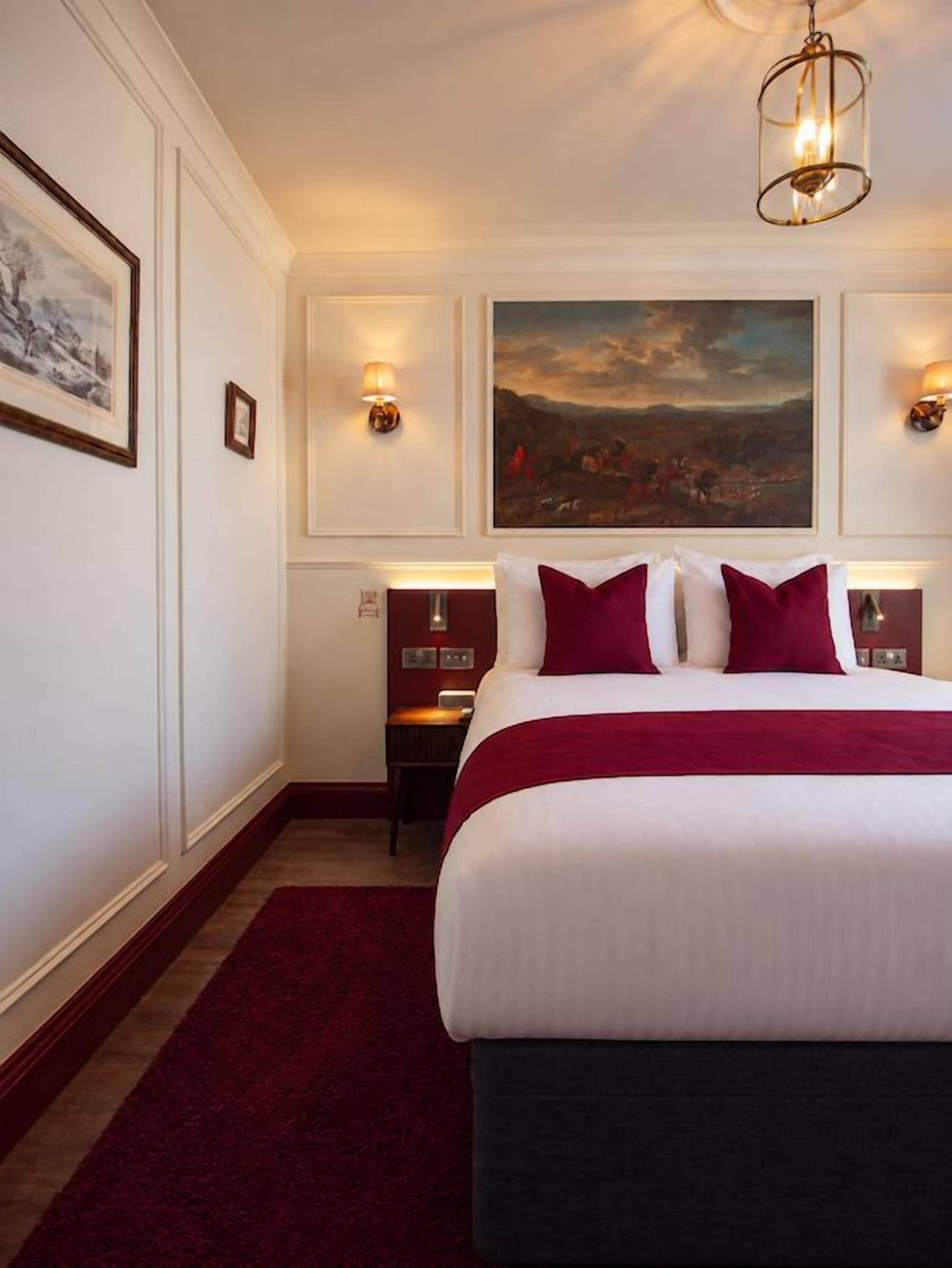 Ein Hotelzimmer mit einem großen Bett, einem roten Teppich, einem Gemälde an der Wand und zwei Wandleuchten.
