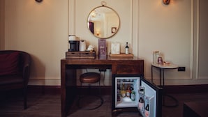 A compact hotel room with a wooden desk, a small refrigerator, a mirror, and two wall-mounted lamps.