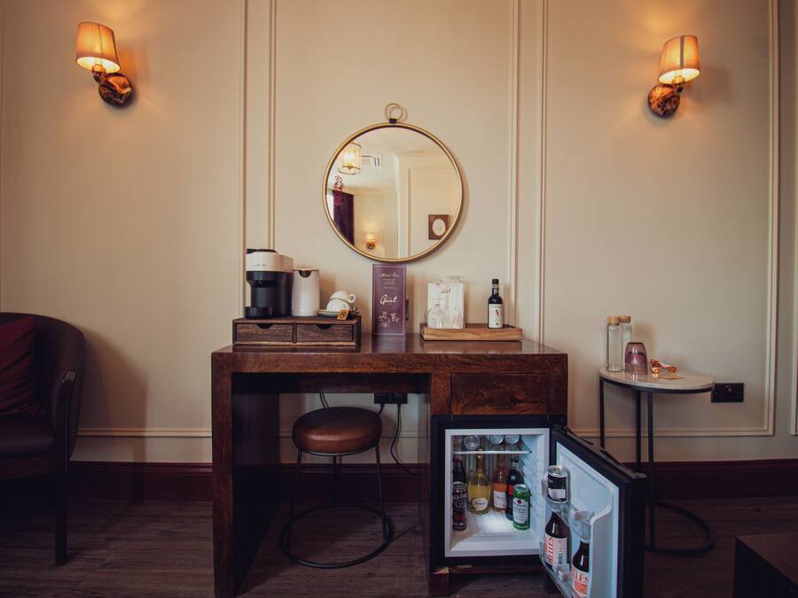 A compact hotel room with a wooden desk, a small refrigerator, a mirror, and two wall-mounted lamps.