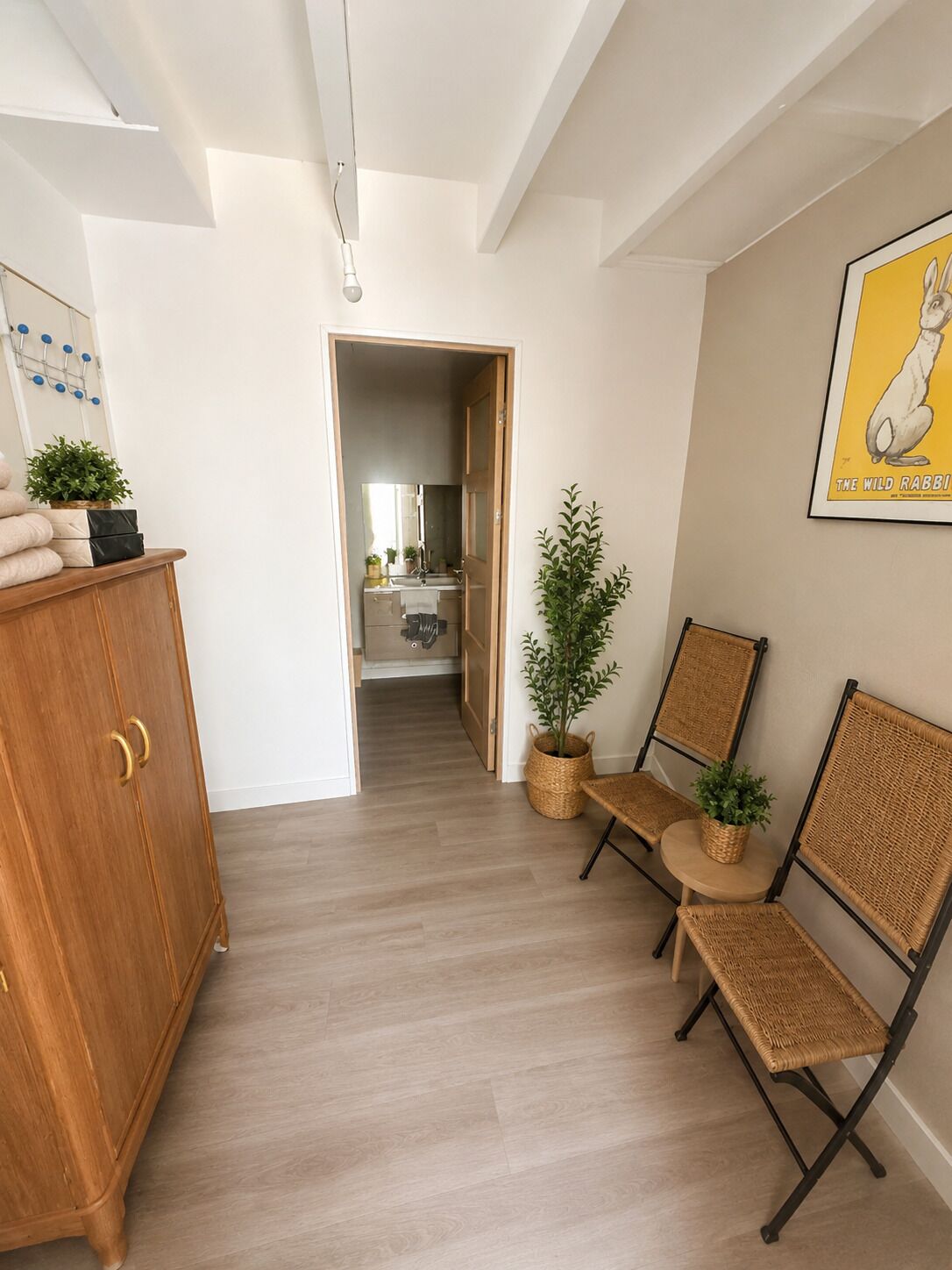 Un couloir avec une armoire en bois, des chaises en osier, une plante en pot et un tableau encadré accroché au mur.