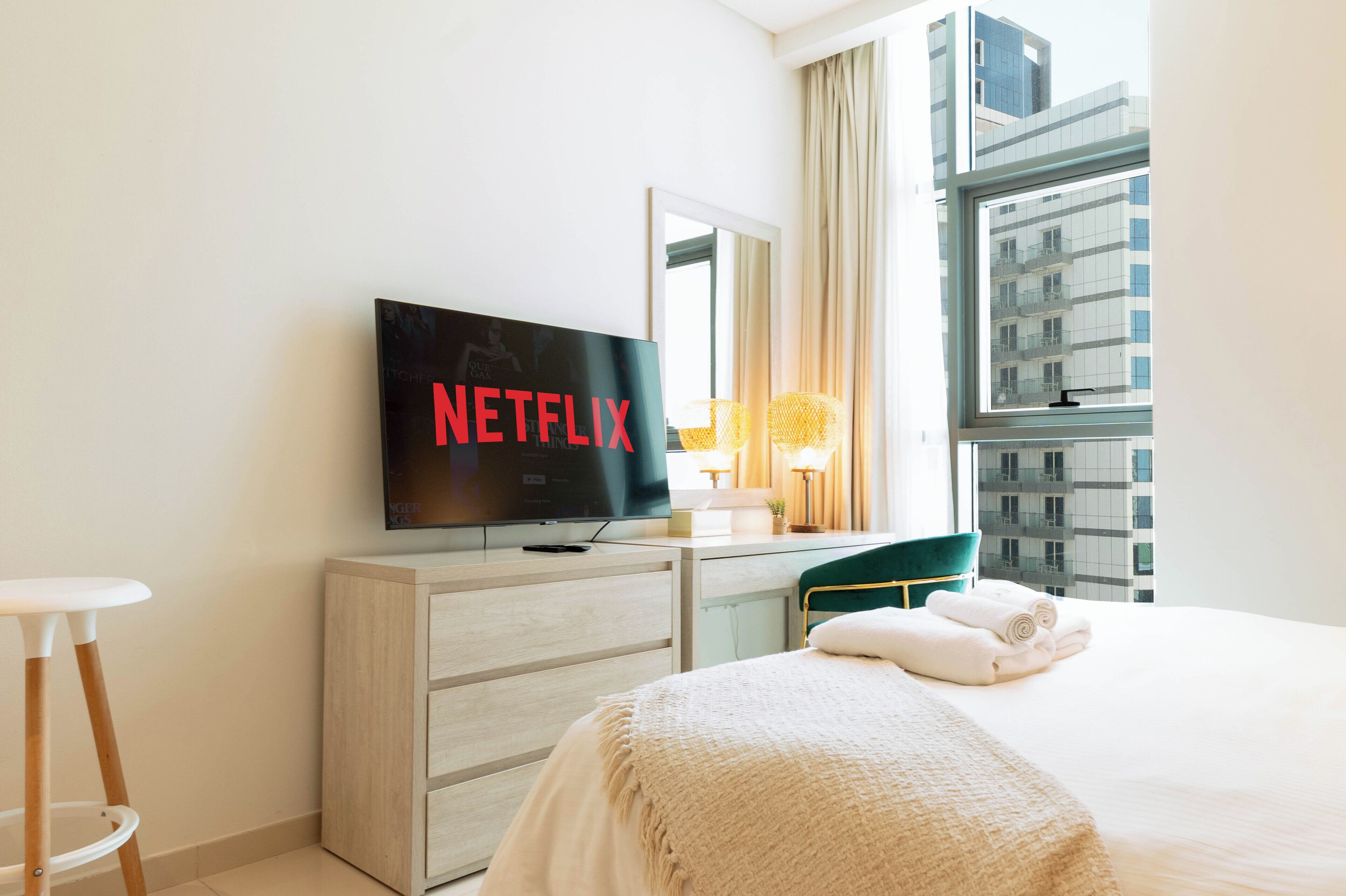 A hotel room with a bed, a TV displaying 'NETFLIX,' a dresser, a window with a view of a building, and a stool.