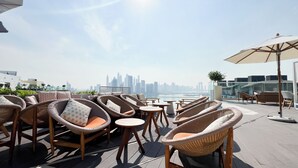 A rooftop terrace with wicker furniture, a city skyline view, and a glass railing. - Elegant Palm Studio with Sea View & Pool (Dubai)
