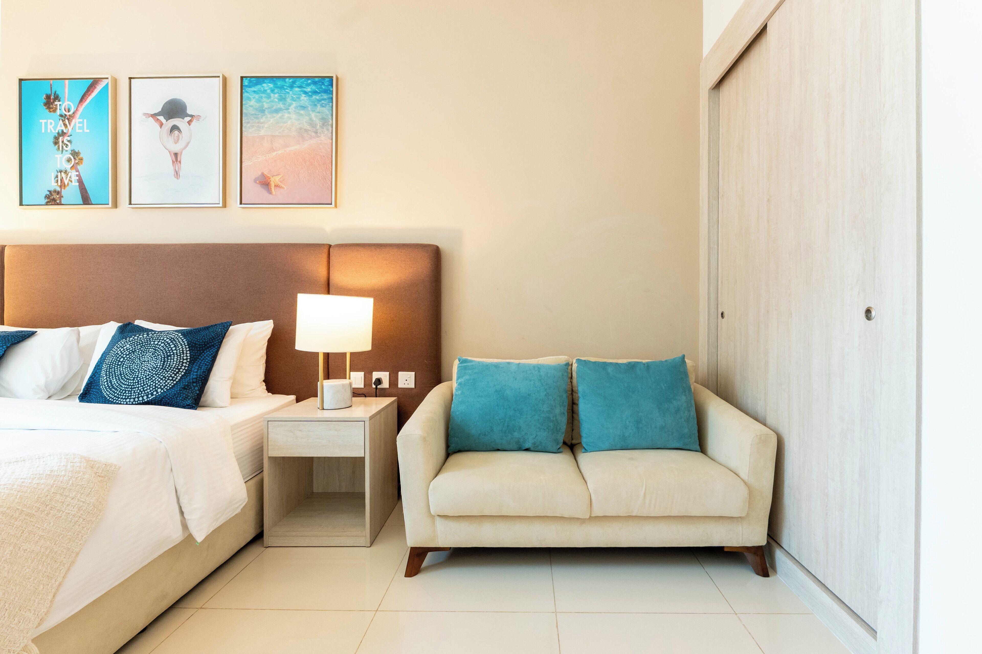 Une chambre d’hôtel avec un lit, une table de chevet avec une lampe, un canapé et trois tableaux encadrés accrochés au mur.