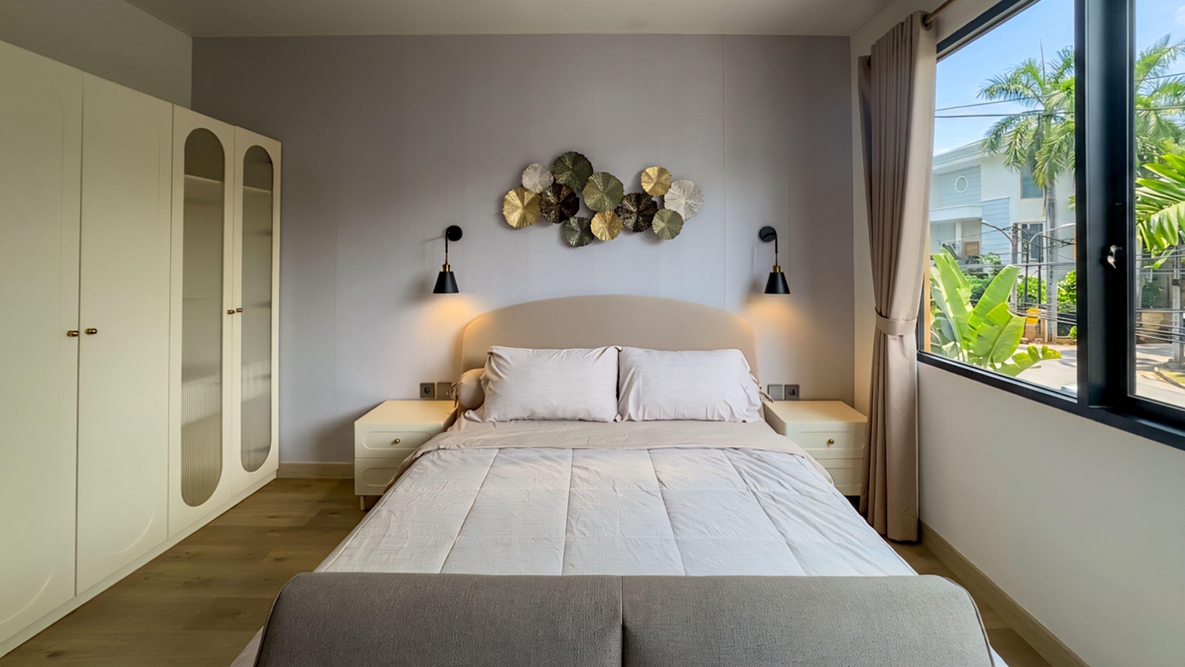 A bedroom with a bed, two bedside tables, a wardrobe, and a window with a view of palm trees.