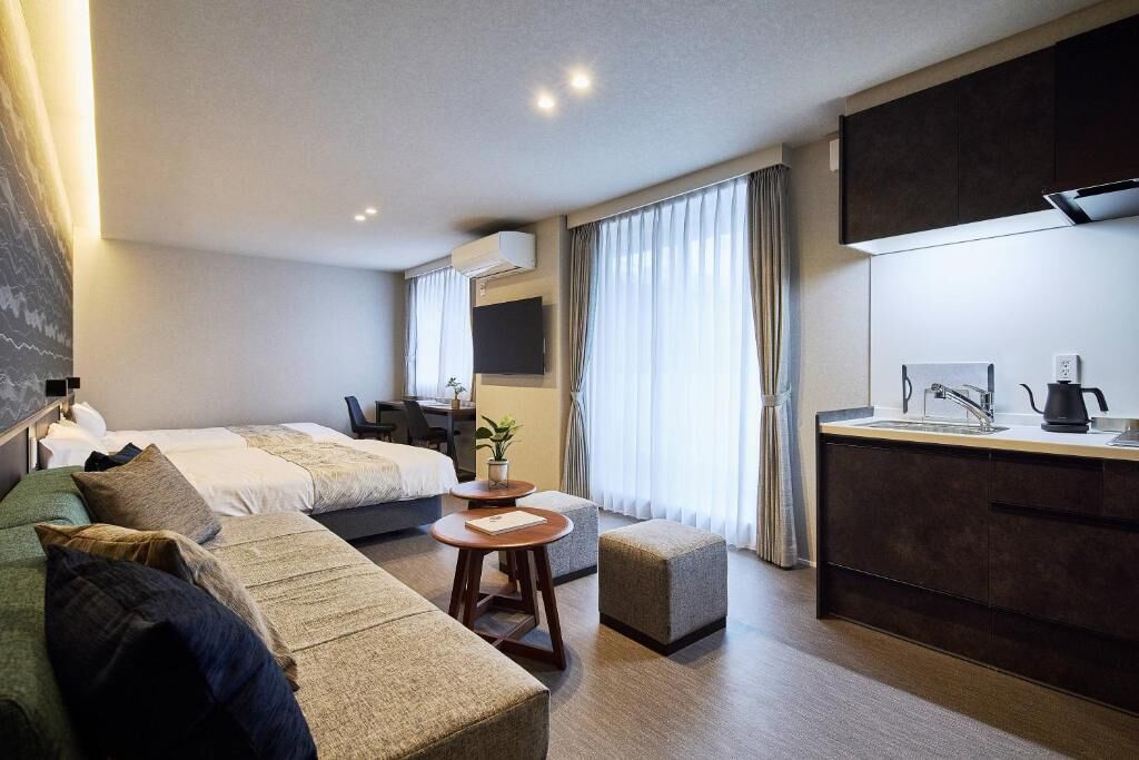 A modern hotel room with a bed, sofa, small table, kitchenette, and a desk.
