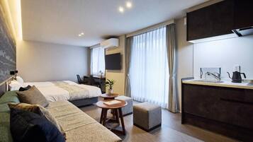 A modern hotel room with a bed, sofa, small table, kitchenette, and a desk.