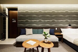A hotel room with a sofa, two round wooden tables, a potted plant, and a bed.